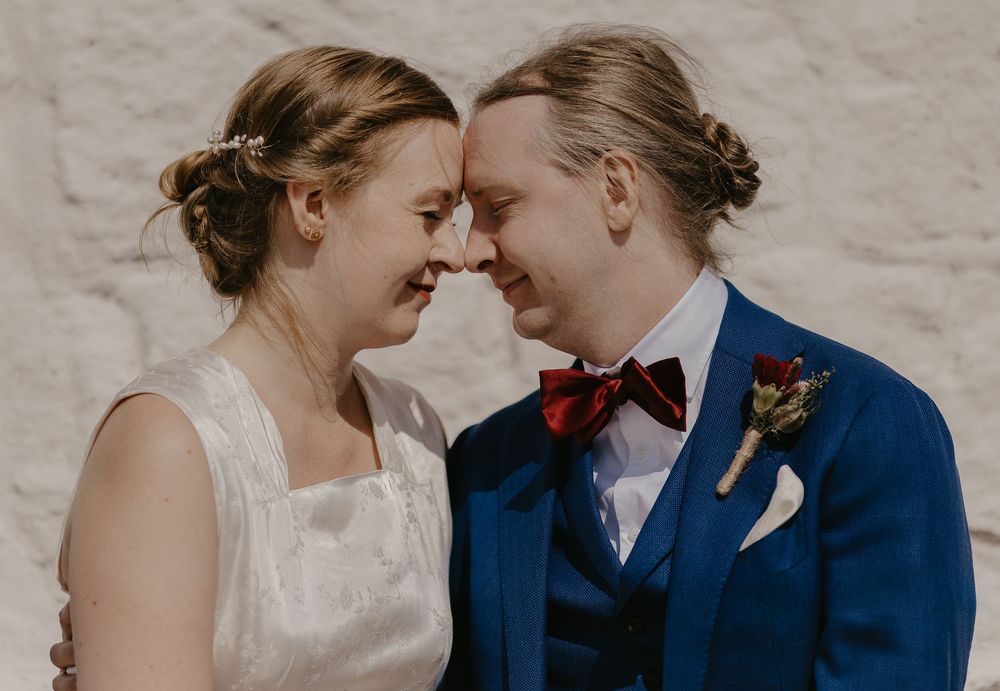 A bride and groom in wedding attire smile warmly at each other, touching foreheads, against a white stone wall.