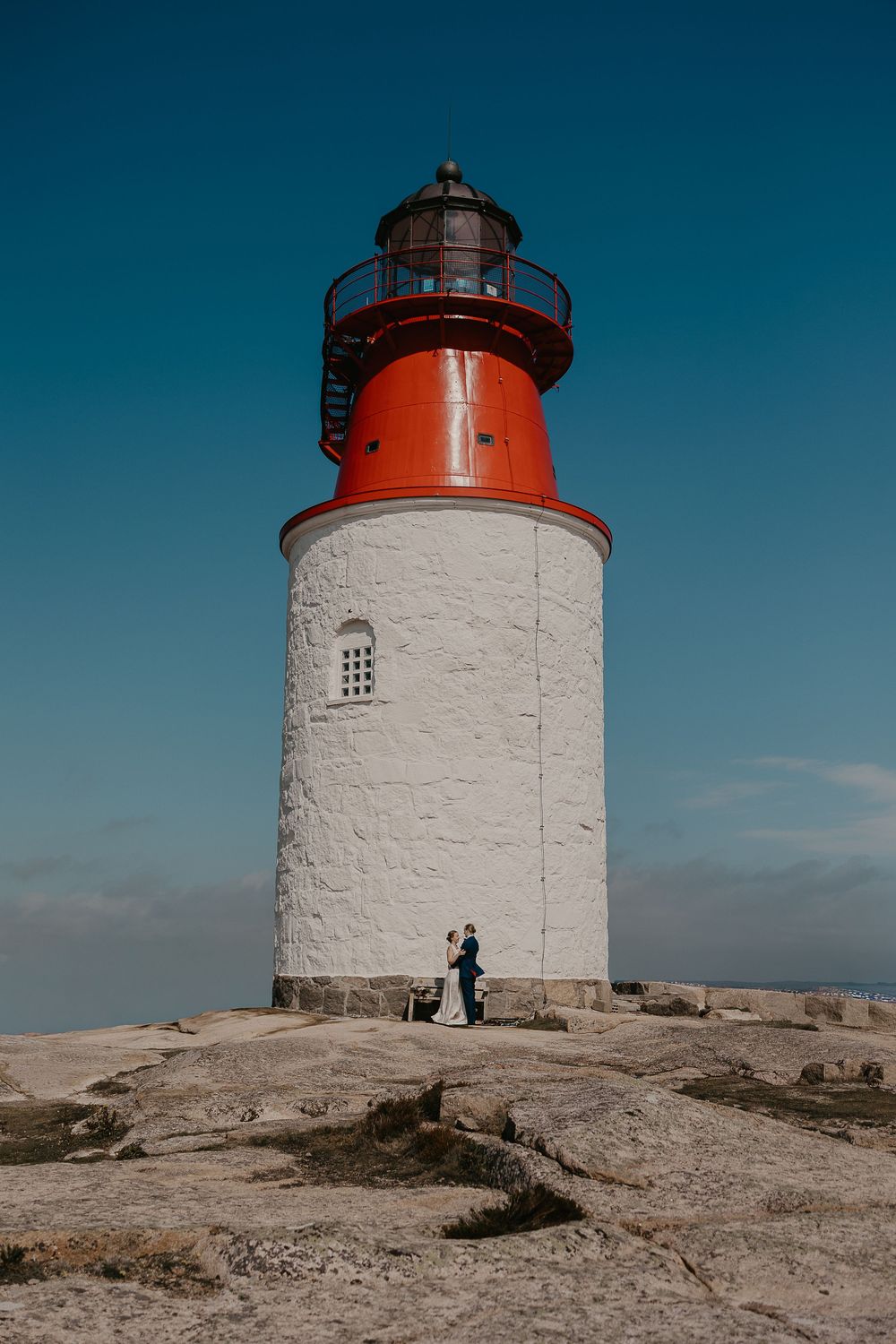Wedding couple embracing at the base of a red and white lighthouse on rocky coastal terrain.