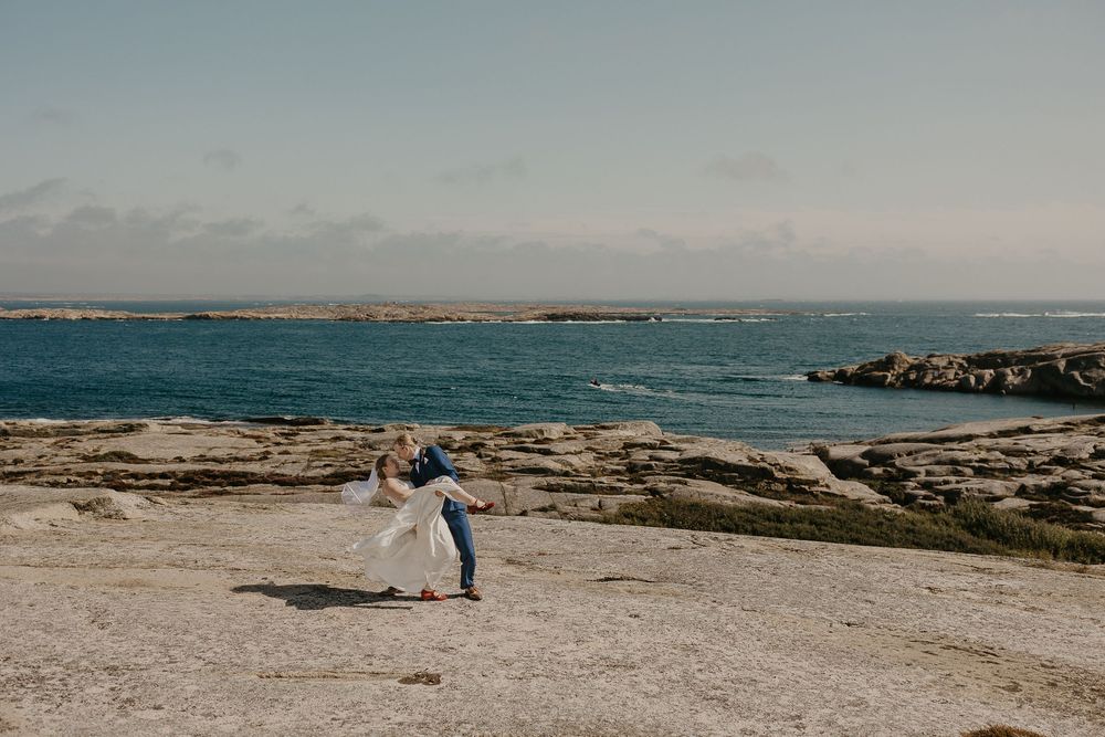 Couple dancing on rocky coastal landscape with turquoise ocean and clear sky in background during wedding photoshoot.
