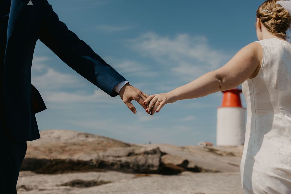 Bride and groom holding hands outdoors near rocky coastline with lighthouse and blue sky on wedding day.