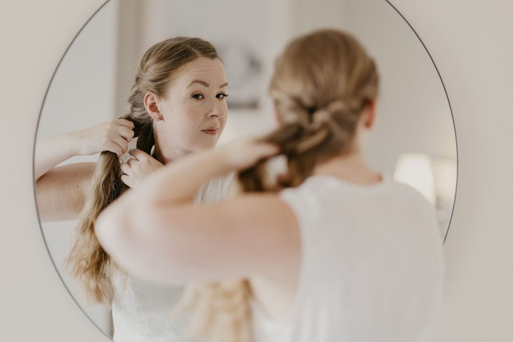 Young woman styling her long hair into a braid while looking at her reflection in a round mirror.