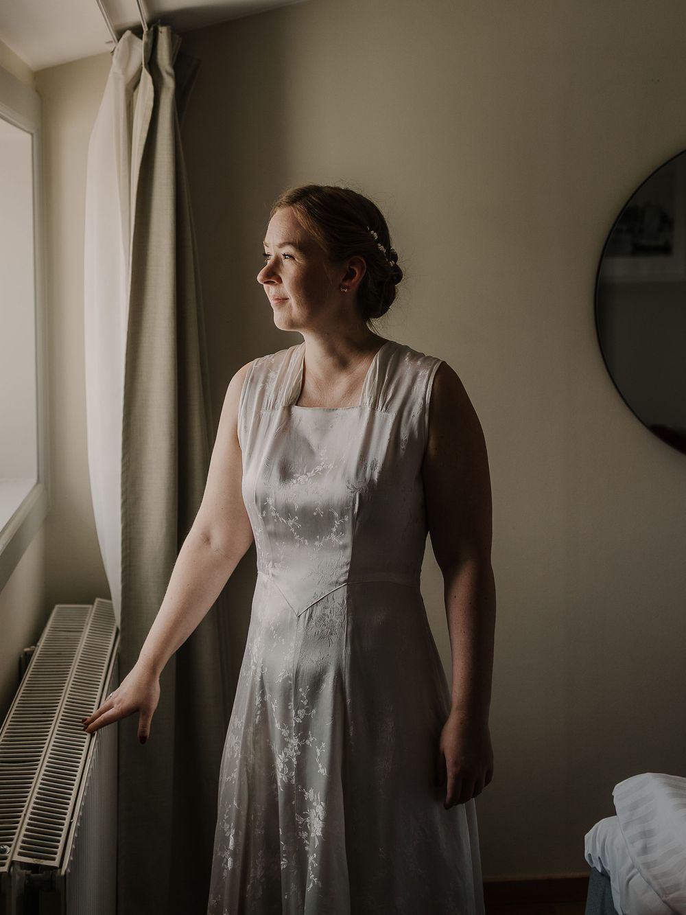 Bride in white lace dress standing by window in soft natural light, looking thoughtfully to the side.