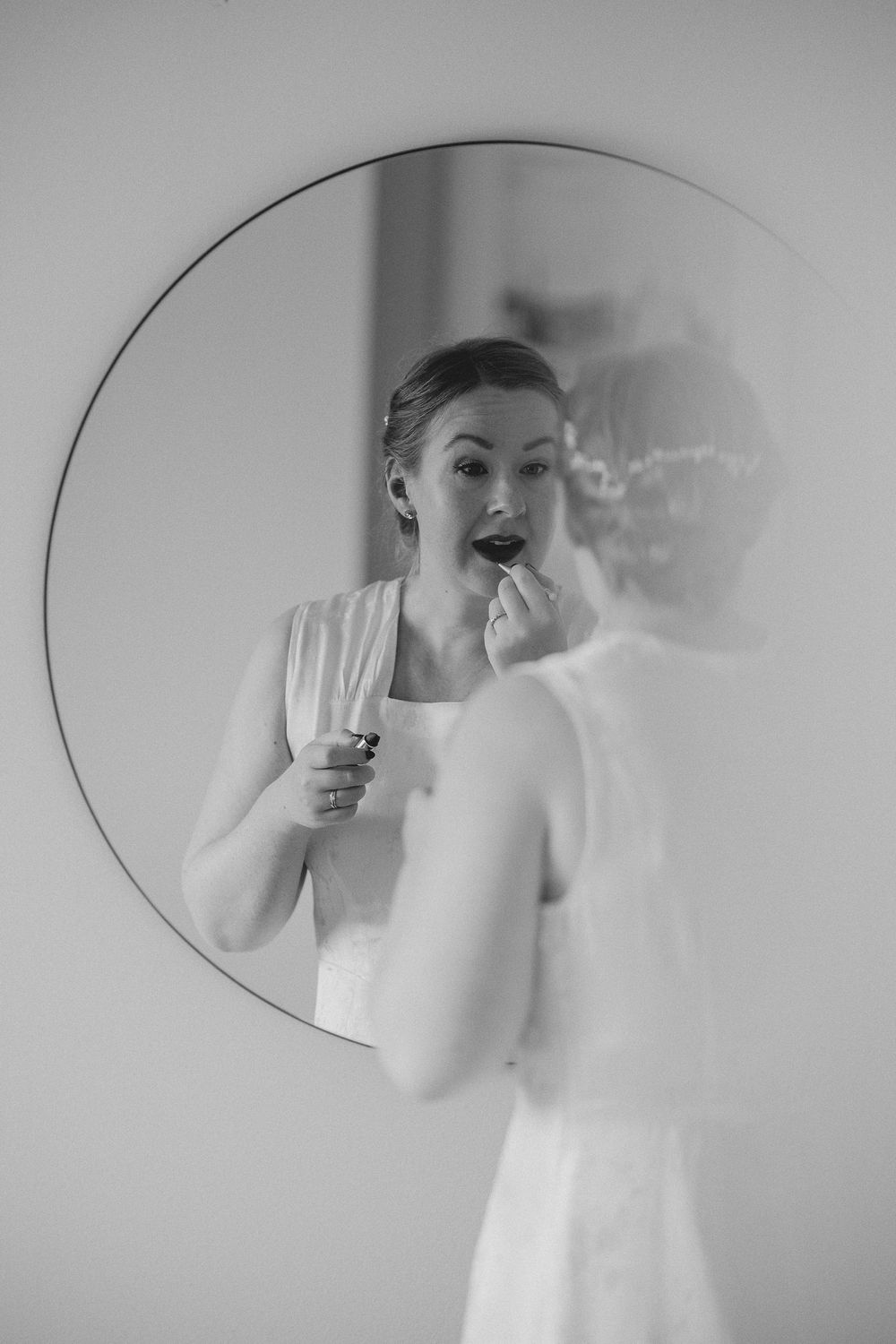 Bride applying lipstick while gazing into a round mirror, captured in elegant black and white photography.