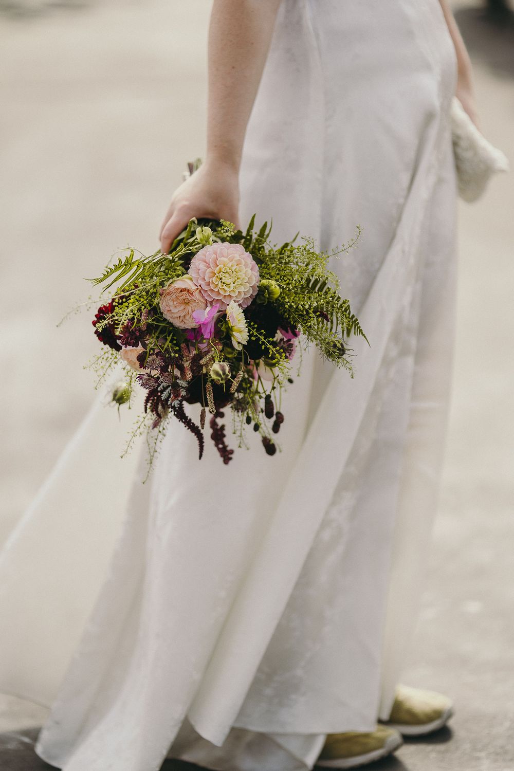 Bride in white gown holding a lush bridal bouquet with pink dahlias, ferns, and trailing greenery.