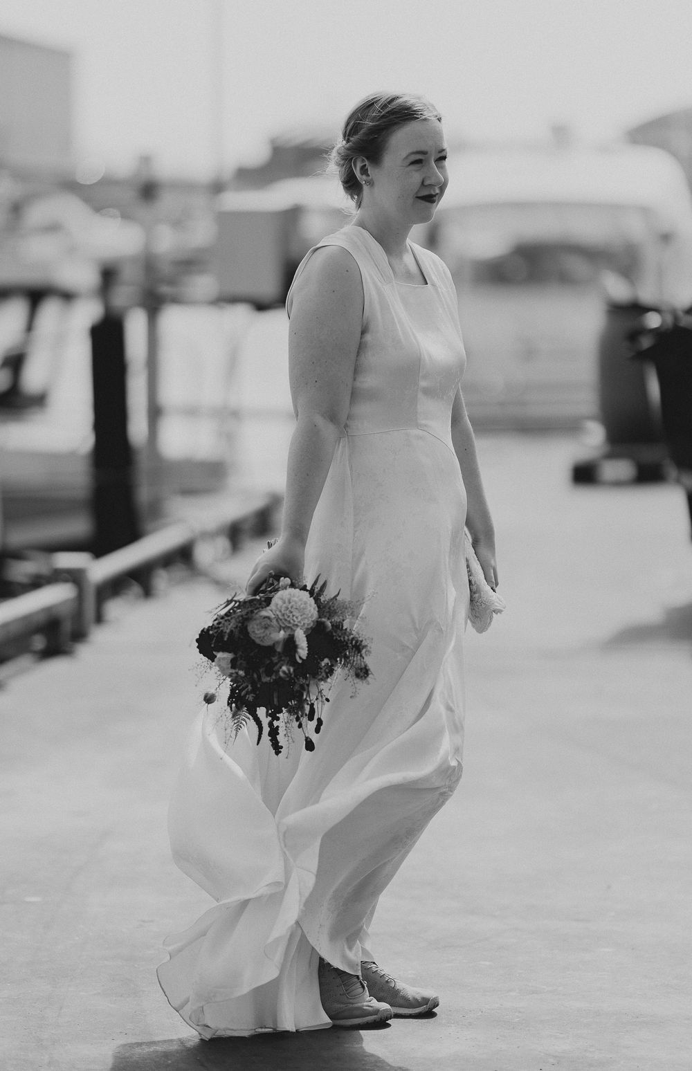 Bride in white gown holding bouquet walks along waterfront dock in black and white wedding photo.