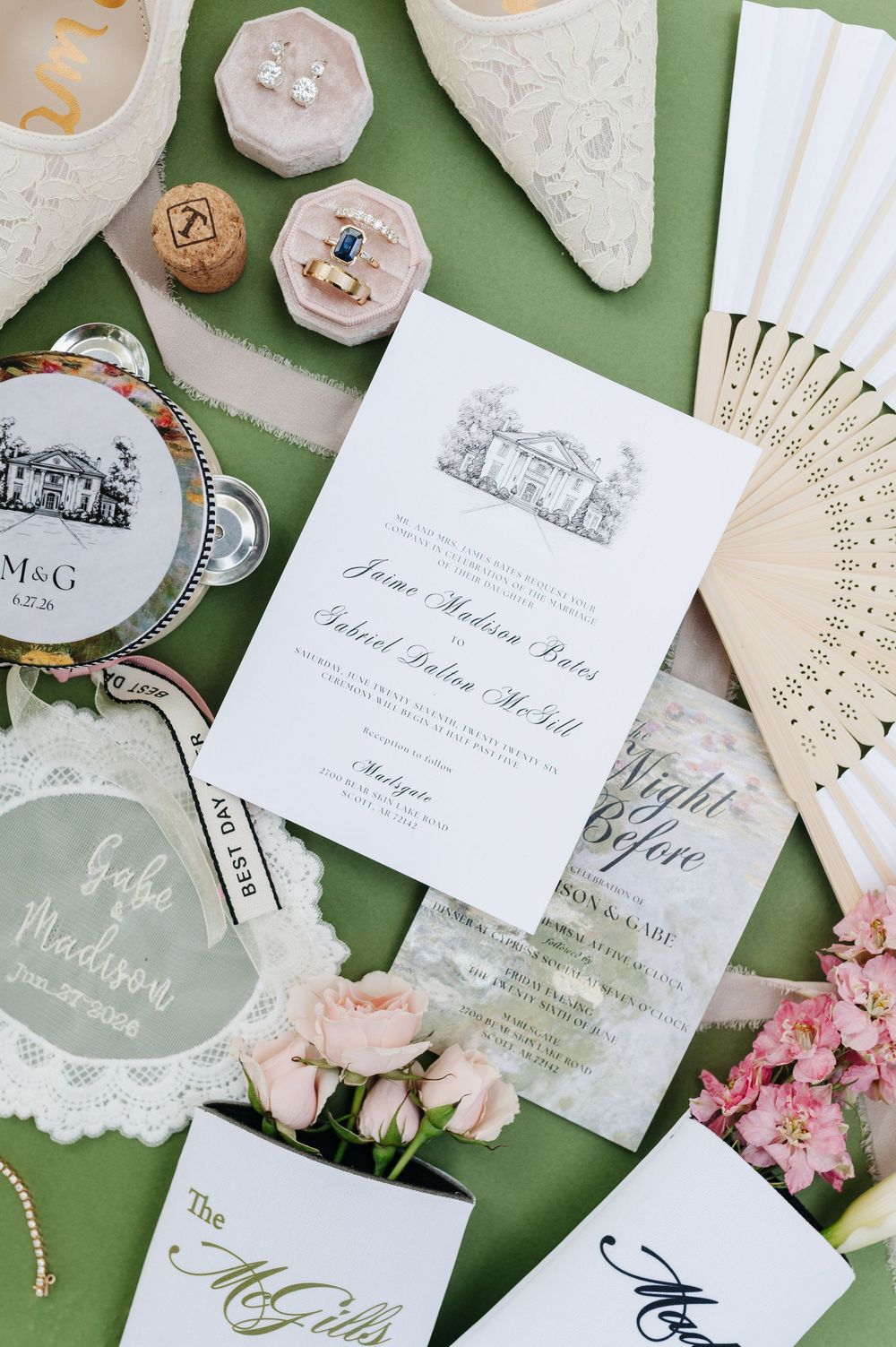 Elegant wedding invitation suite with floral accents, rings, and accessories on a green background.