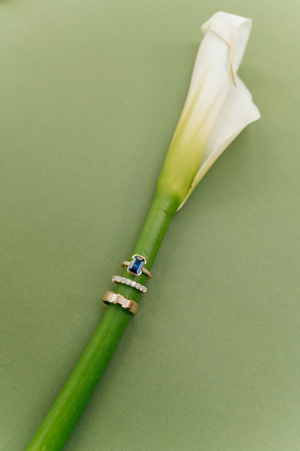 Gold wedding bands and sapphire engagement ring displayed on a white calla lily stem against green background.
