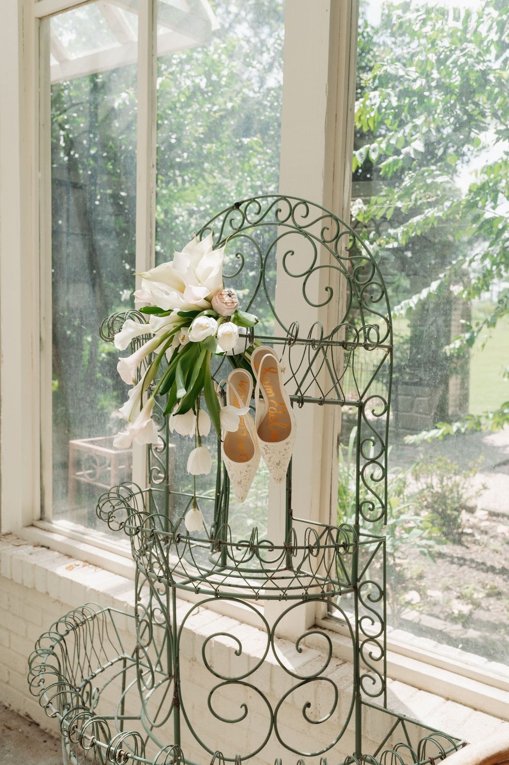 Elegant bridal bouquet and lace heels displayed on ornate green wrought iron chair by sunlit window.