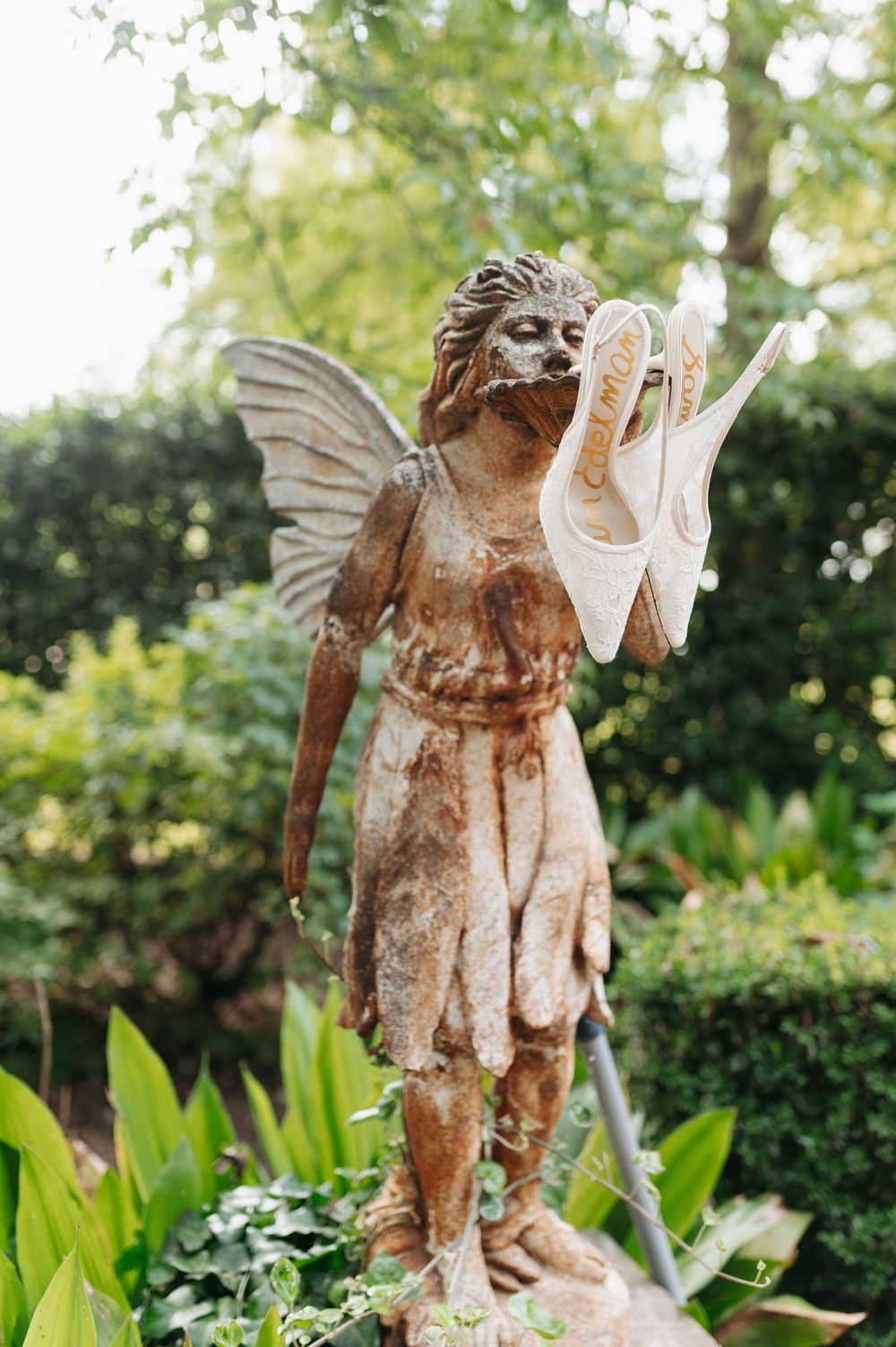 Weathered fairy garden statue holding white bridal shoes surrounded by lush green foliage outdoors.