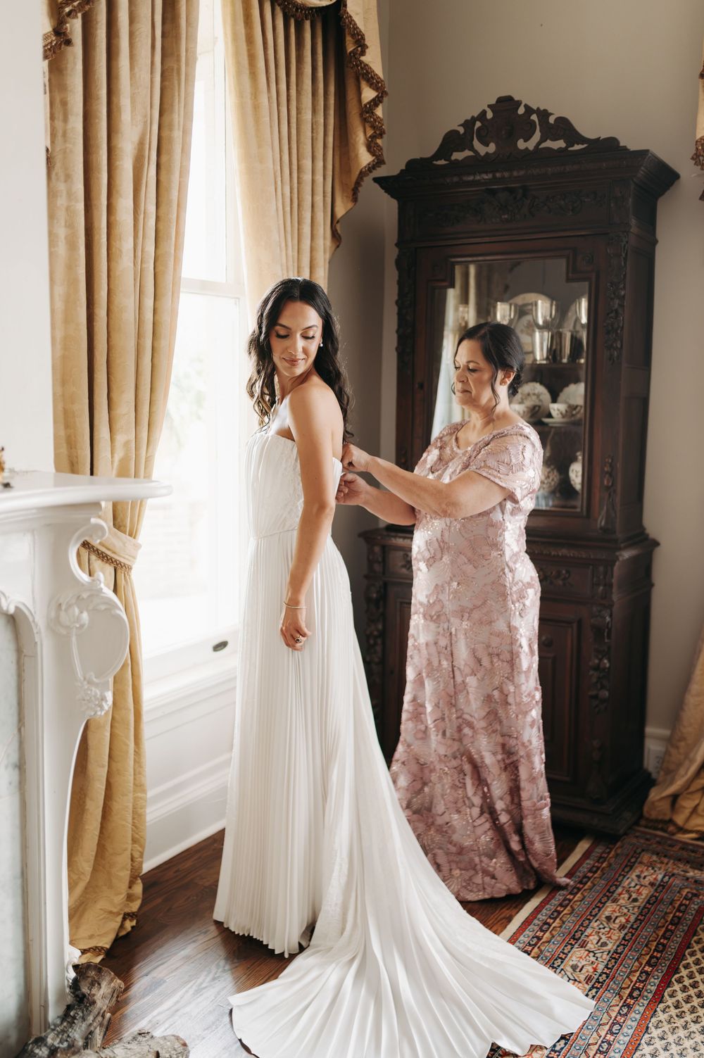 Bride in white pleated gown being helped by woman in pink floral dress near antique cabinet and window.