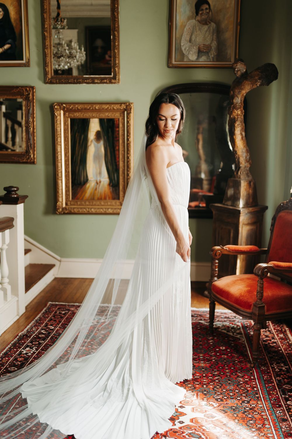 Bride in a white pleated gown with a long veil stands in an elegant room adorned with gold-framed paintings.