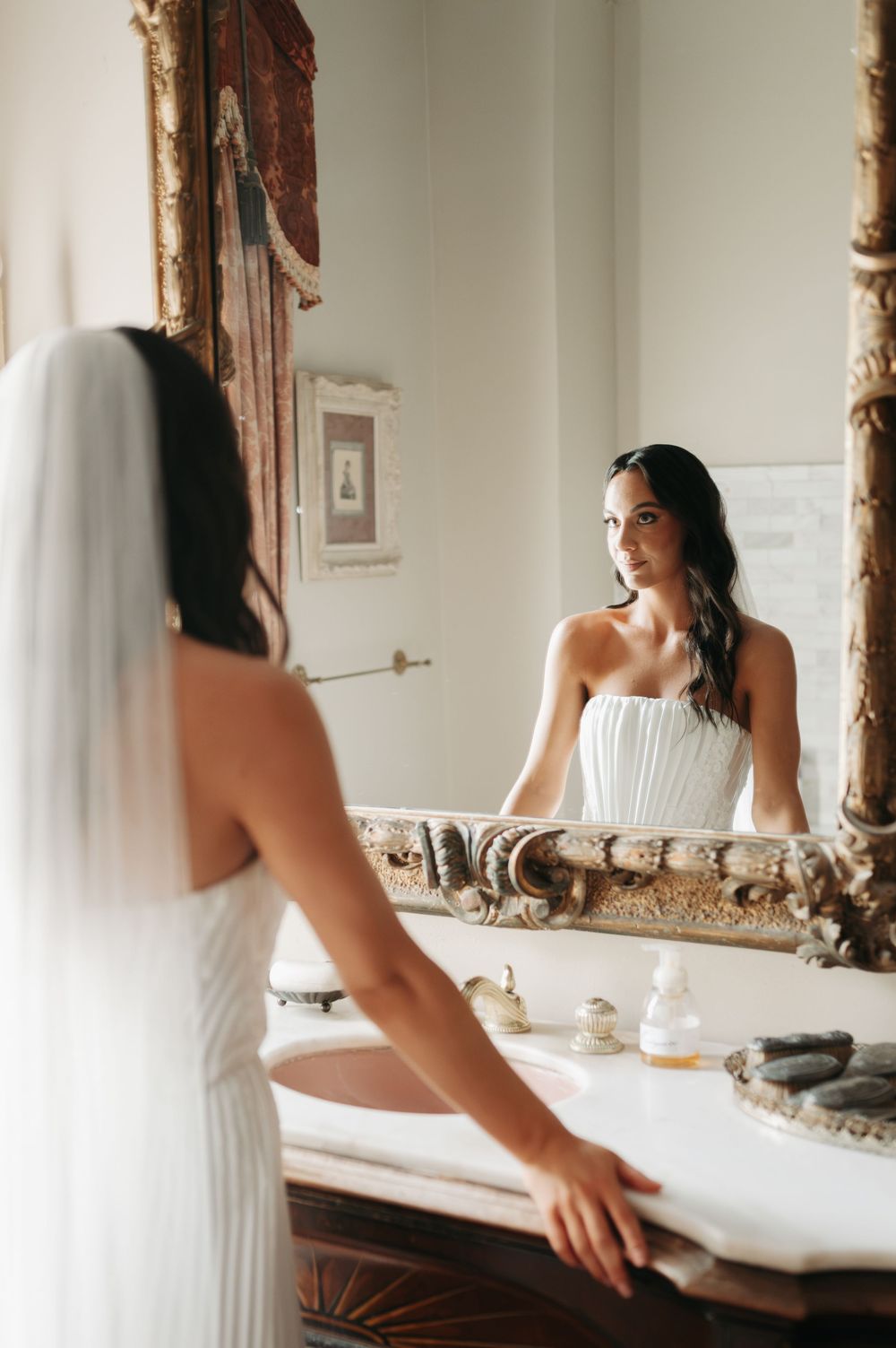 Bride in white strapless gown with veil gazes at her reflection in an ornate gold-framed bathroom mirror.