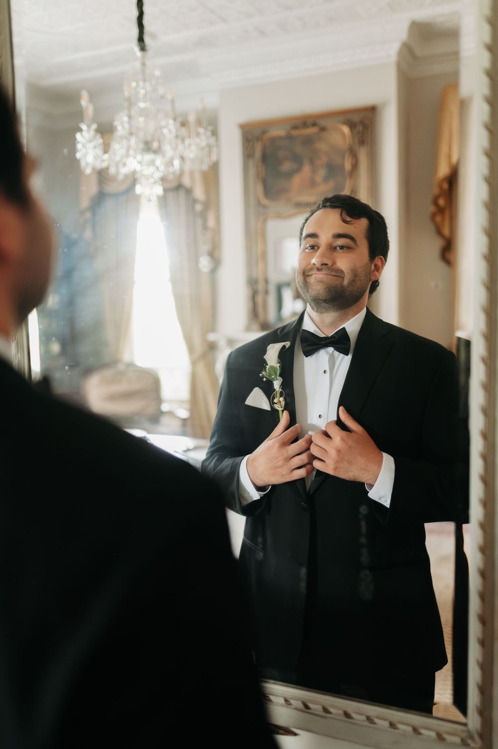 Groom in black tuxedo with boutonniere adjusts jacket while smiling at his reflection in an ornate mirror.