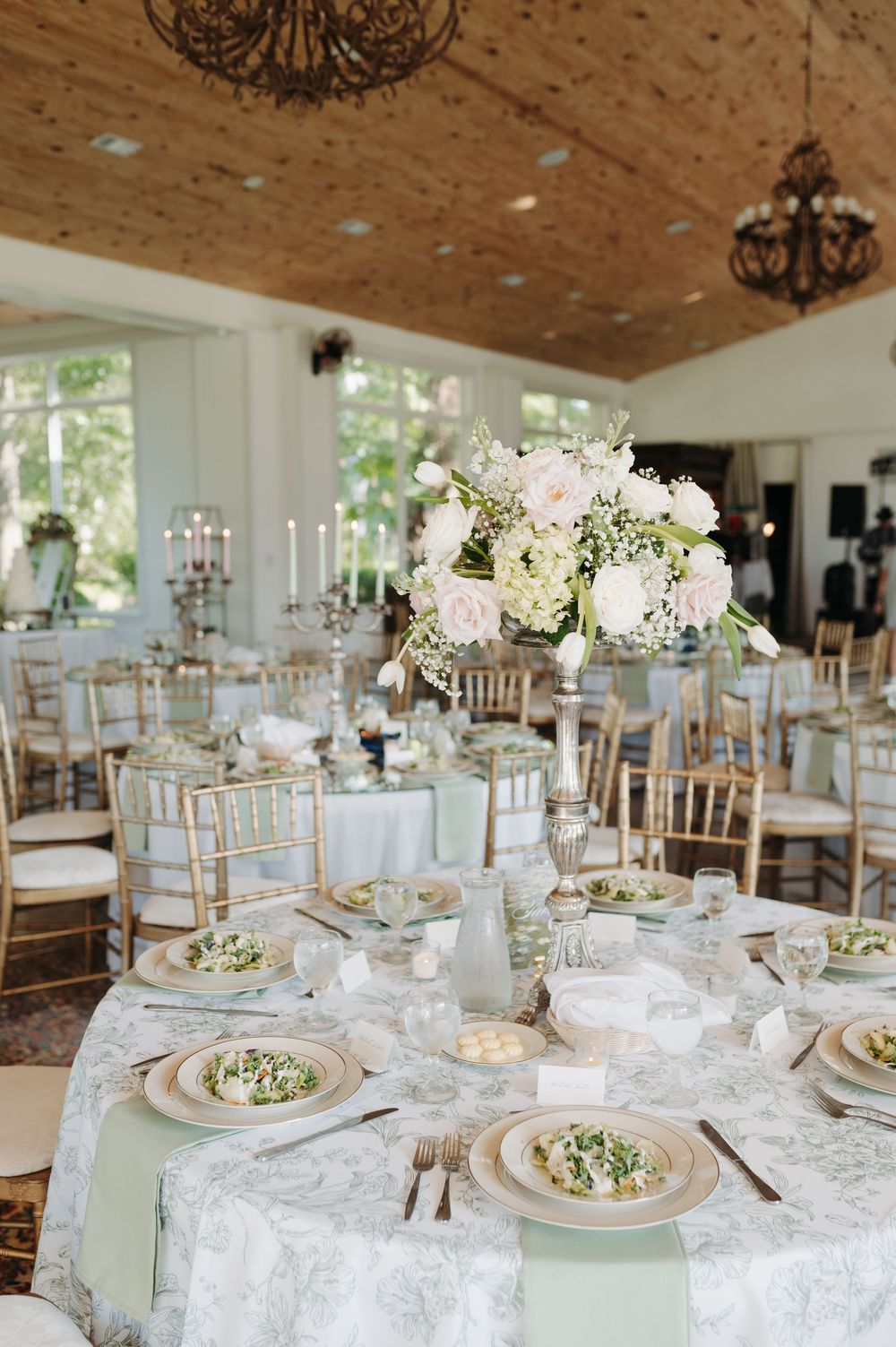 Elegant wedding reception table with blush floral centerpiece, gold chiavari chairs, and sage green napkins on damask linens.