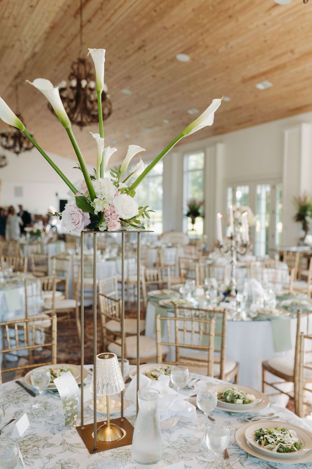 Elegant wedding reception with tall gold centerpieces featuring white calla lilies and pink roses on floral tablecloths.