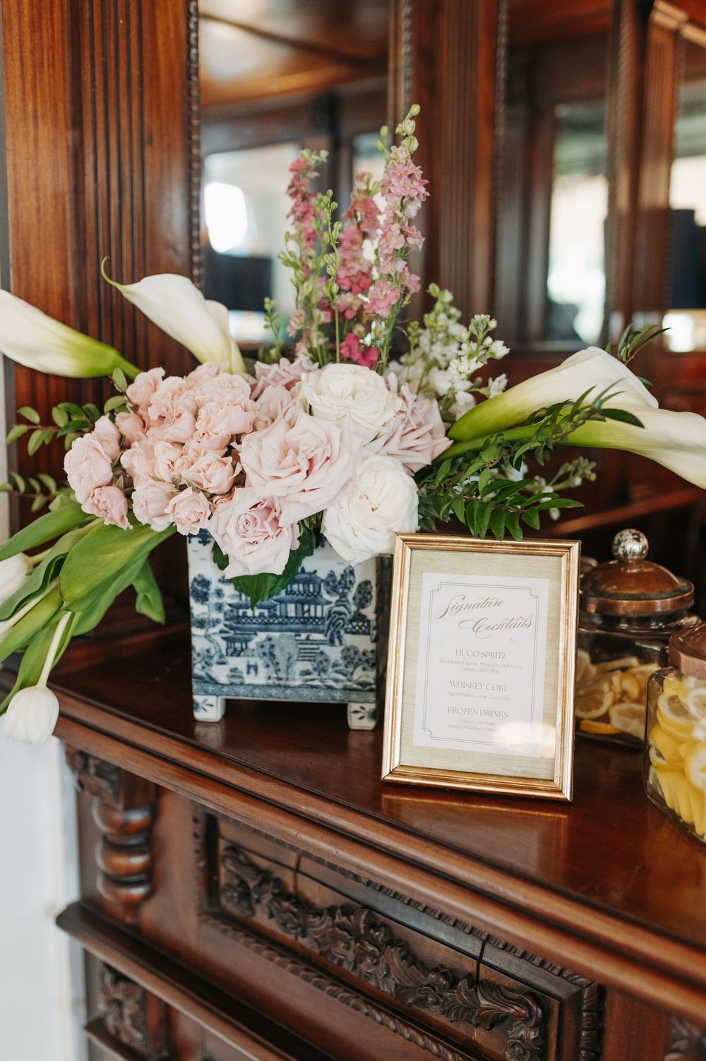 Blue and white chinoiserie vase with blush roses, calla lilies, and pink florals beside a gold-framed menu card on a mahogany dresser.