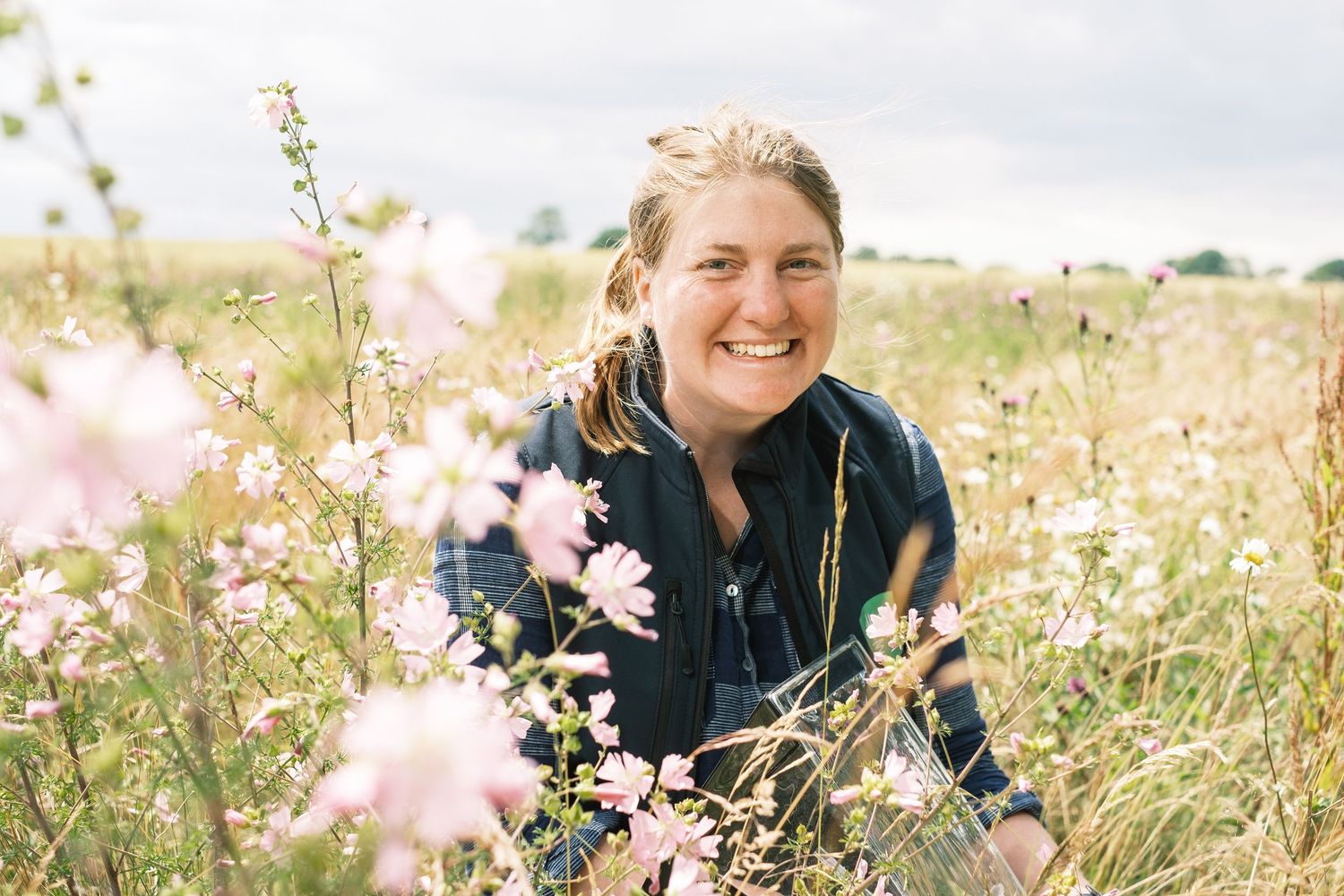 Suffolk Wildlife Trust. A smiling woman surrounded by pink wildflowers in a meadow field on a cloudy day. Photographed by John Ferguson