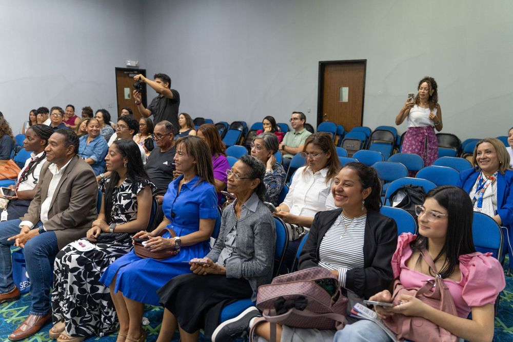 Diverse audience seated in blue chairs at a conference or seminar in a modern meeting room.