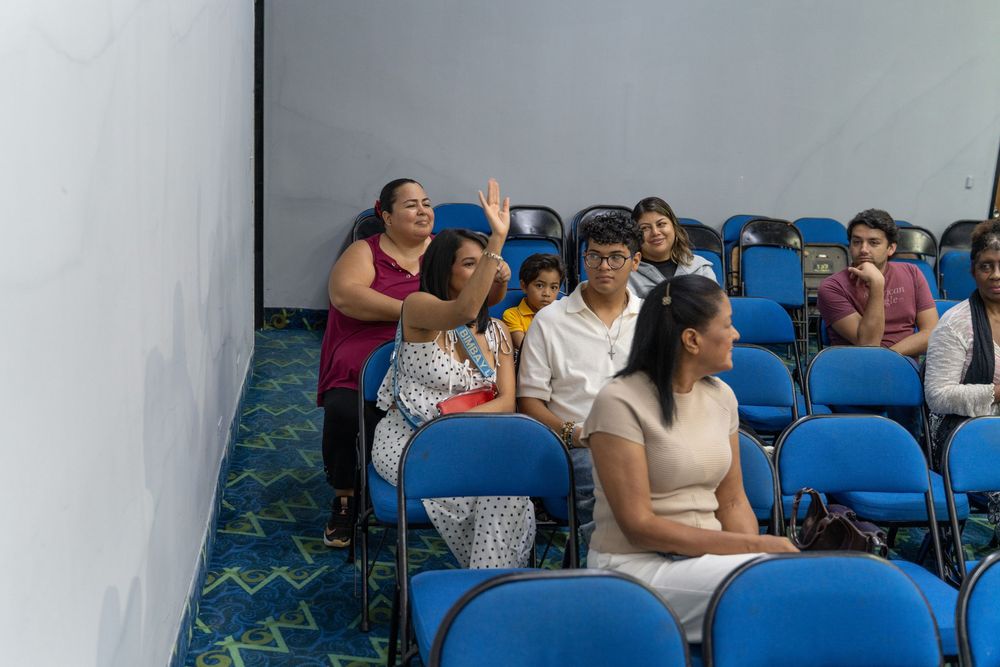 People sitting in blue chairs in a small auditorium, with a woman raising her hand during a presentation.