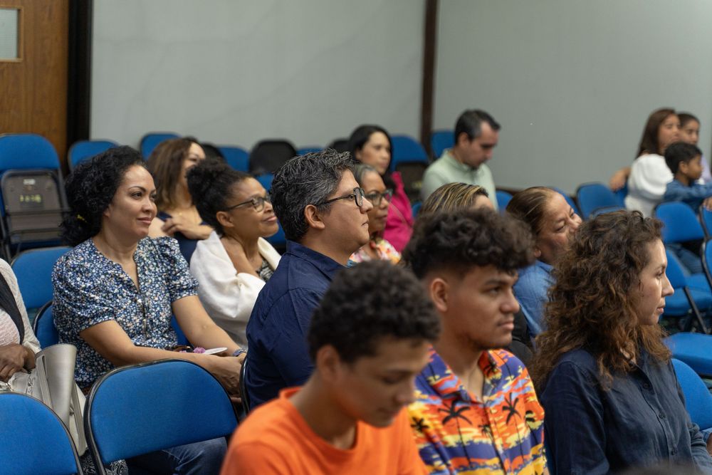 Diverse audience seated in blue chairs at an indoor lecture or seminar, engaged and attentive in a modern conference room.