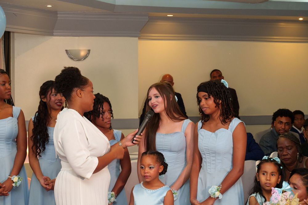 Bridesmaids in light blue dresses gather during a wedding ceremony while a woman in white speaks to the group.