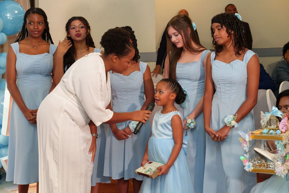 Women and girls in light blue dresses at a formal celebration, with one woman leaning down to speak to a young girl.