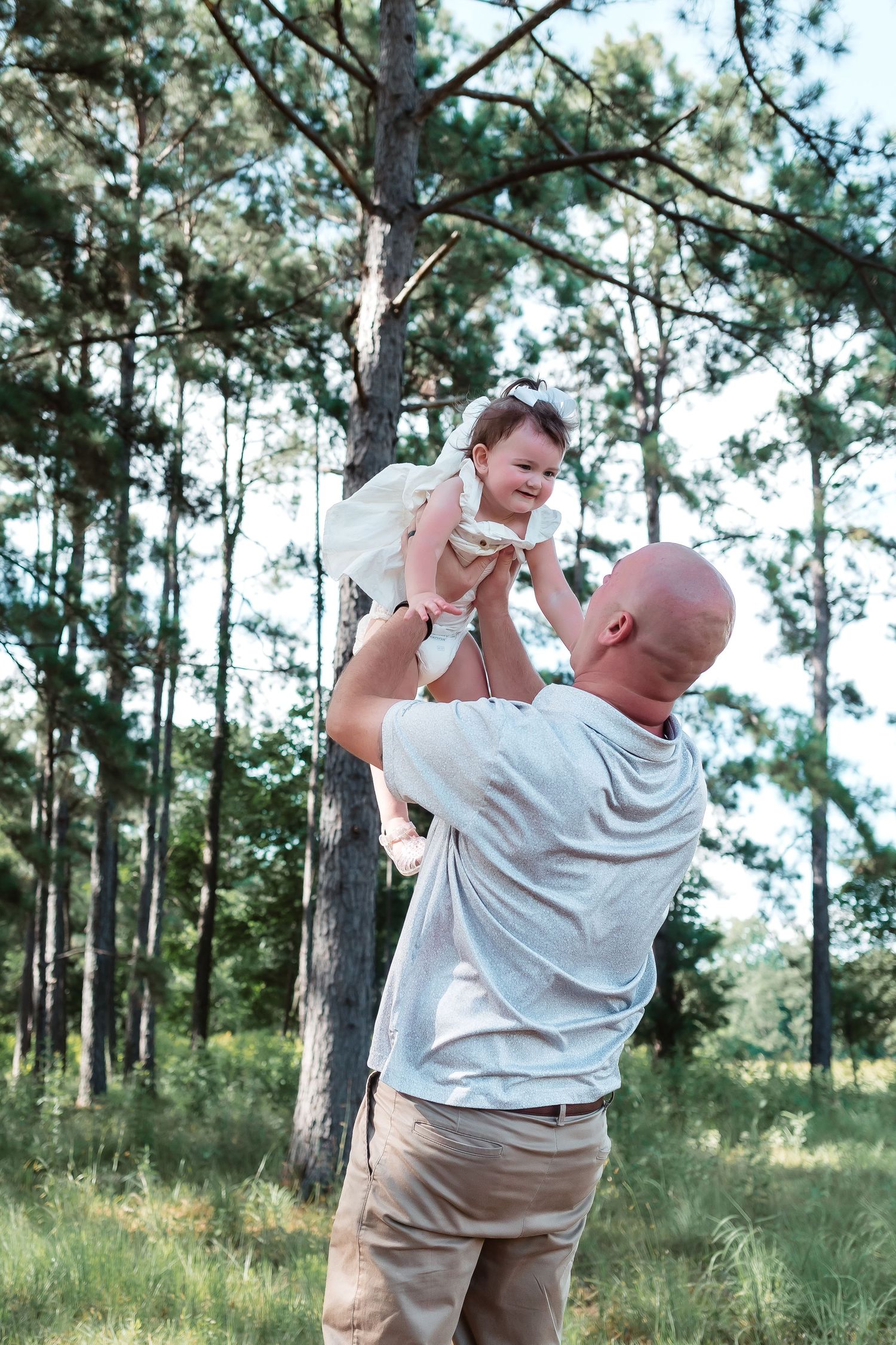 A bald man lifts a smiling baby girl in a white dress above his head in a lush green pine forest.