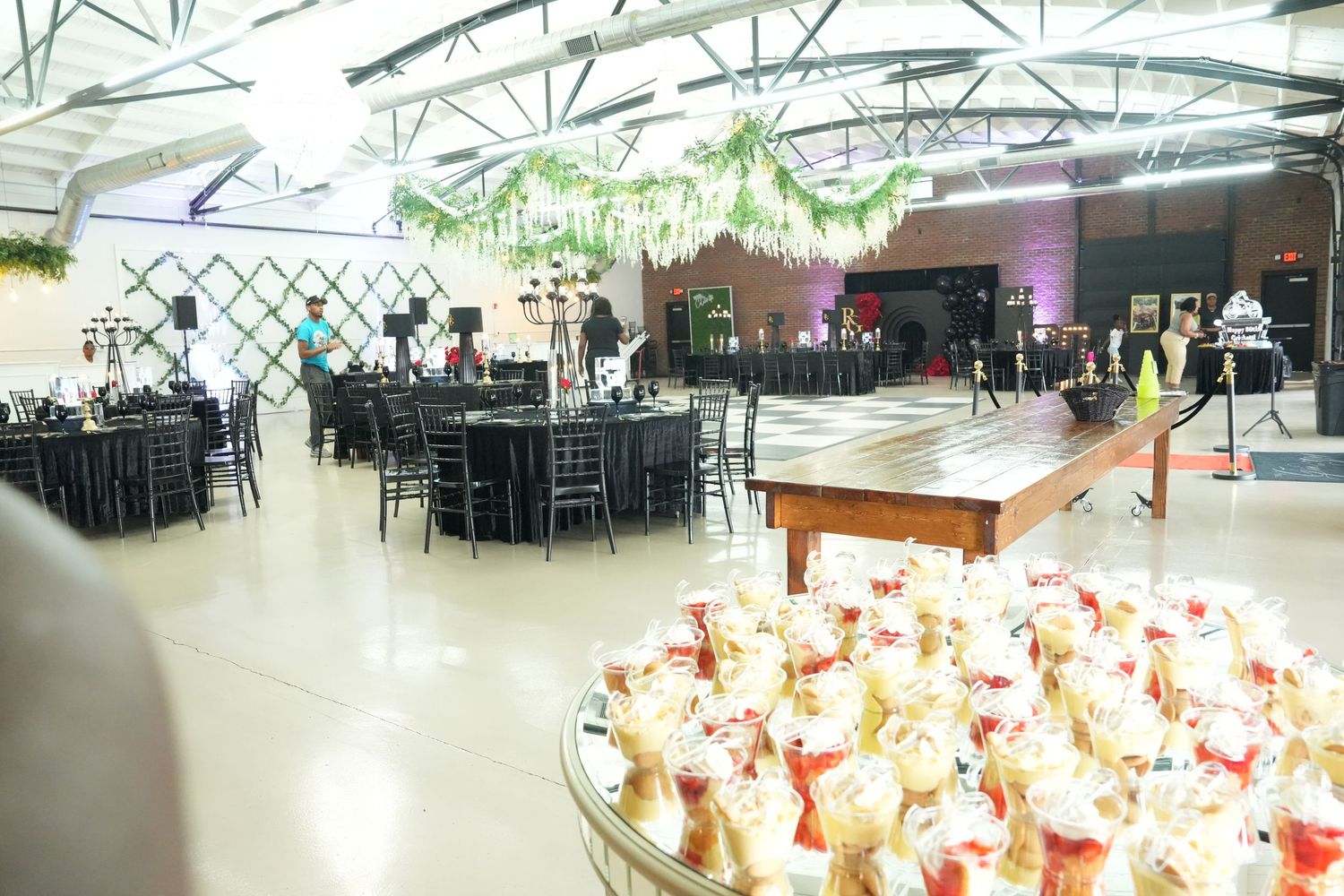 Elegant event venue with black chiavari chairs, dance floor, hanging greenery, and a foreground display of champagne bottles.