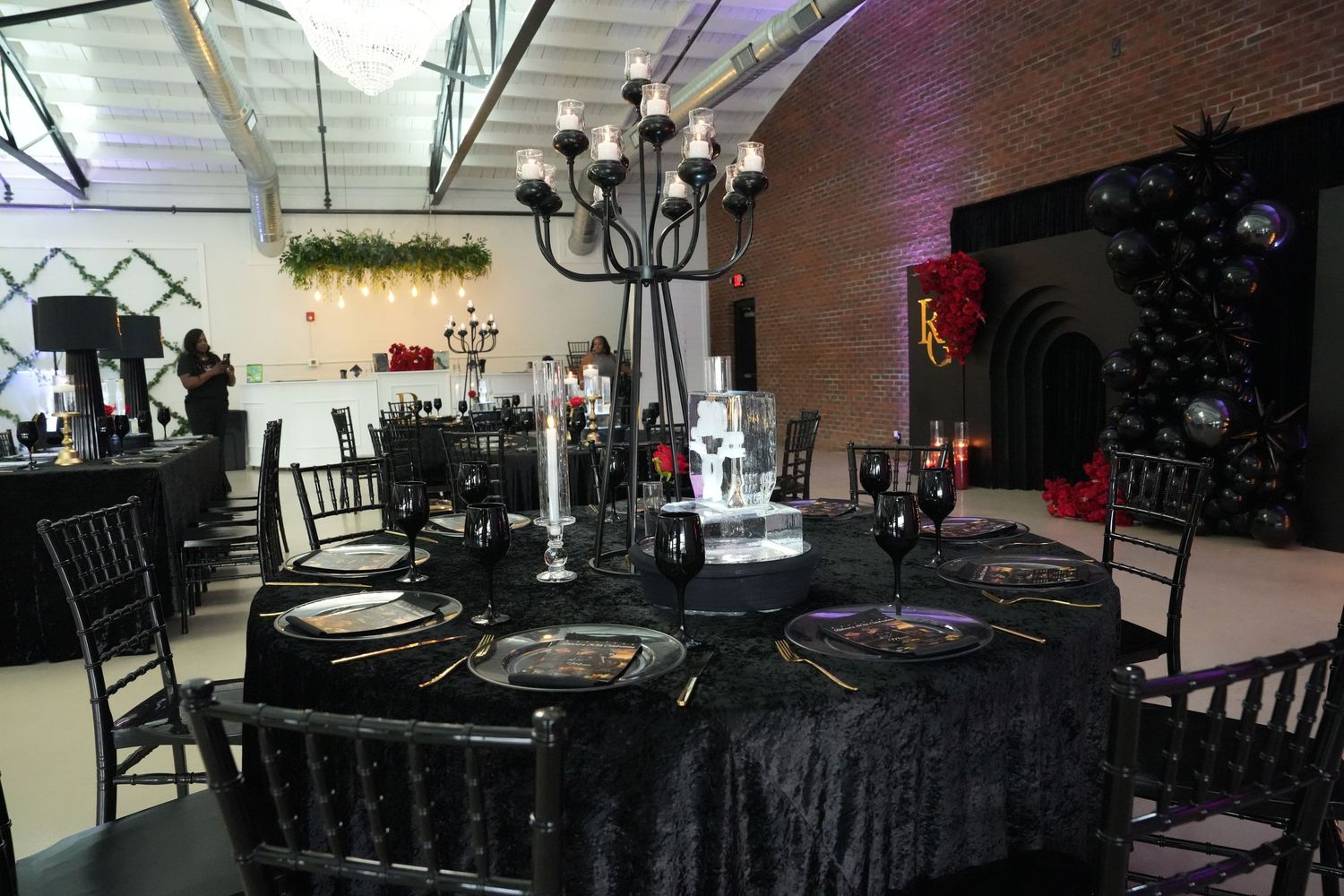 Elegant black-themed event space with chandelier, draped tables, gold accents, and dark balloon arch decor.