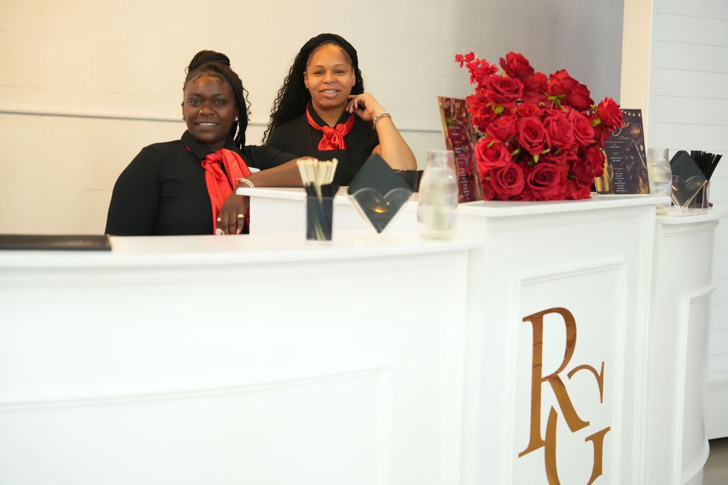 Two smiling women in black attire stand behind a white reception desk adorned with red roses and RCG logo.