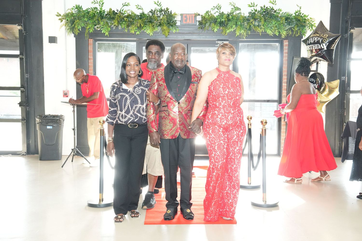 Three people posing on a red carpet at a formal event, dressed in elegant red and black outfits.