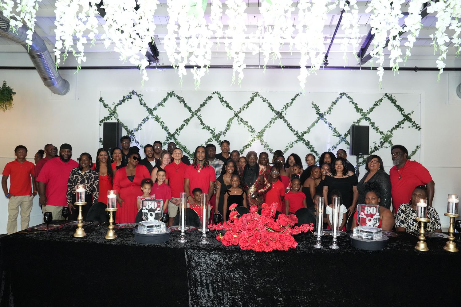 Large group in red and black attire posing together at an elegant event with floral decorations and trophies on display.