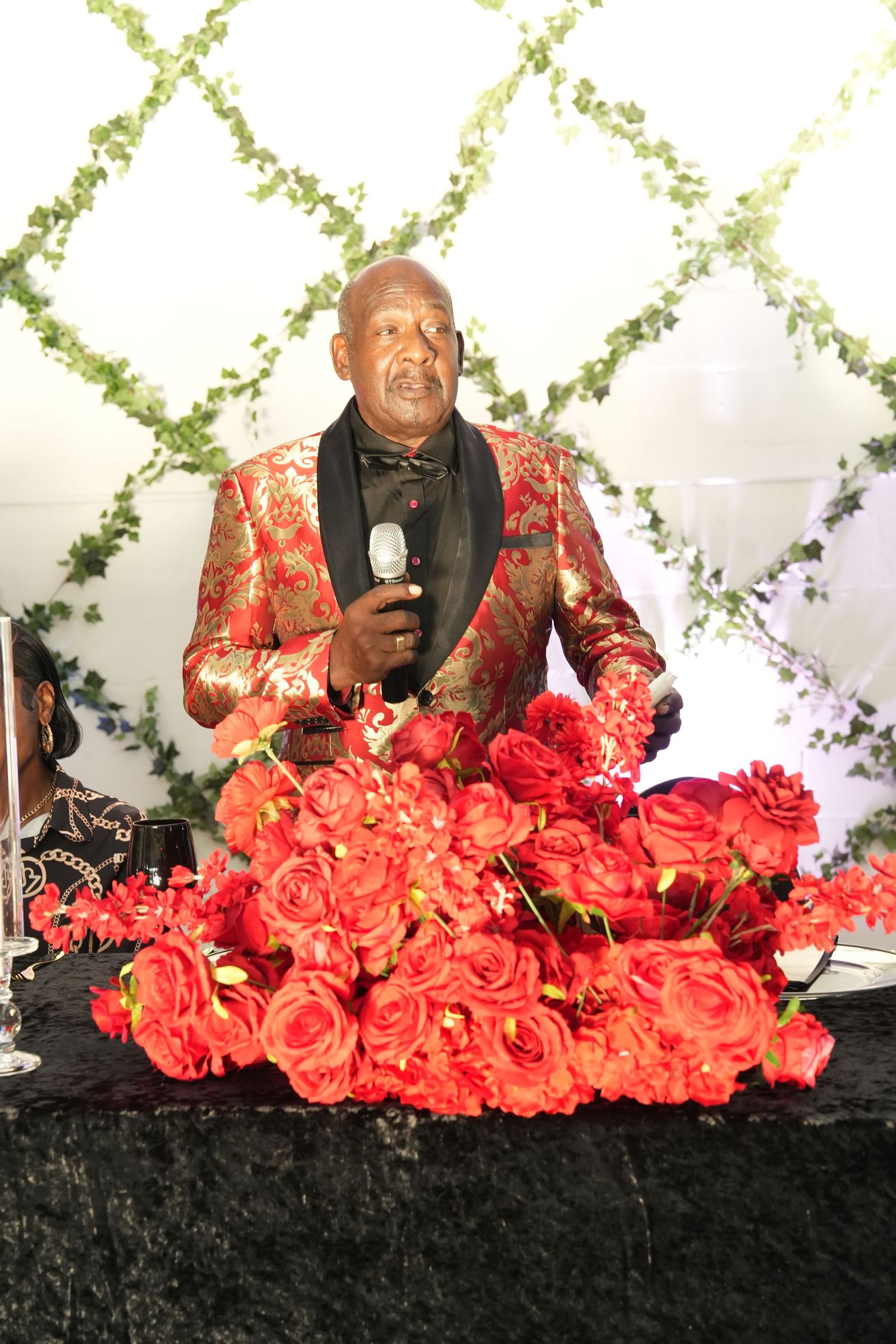 Elderly man in red floral blazer speaking into microphone surrounded by red roses at elegant event.