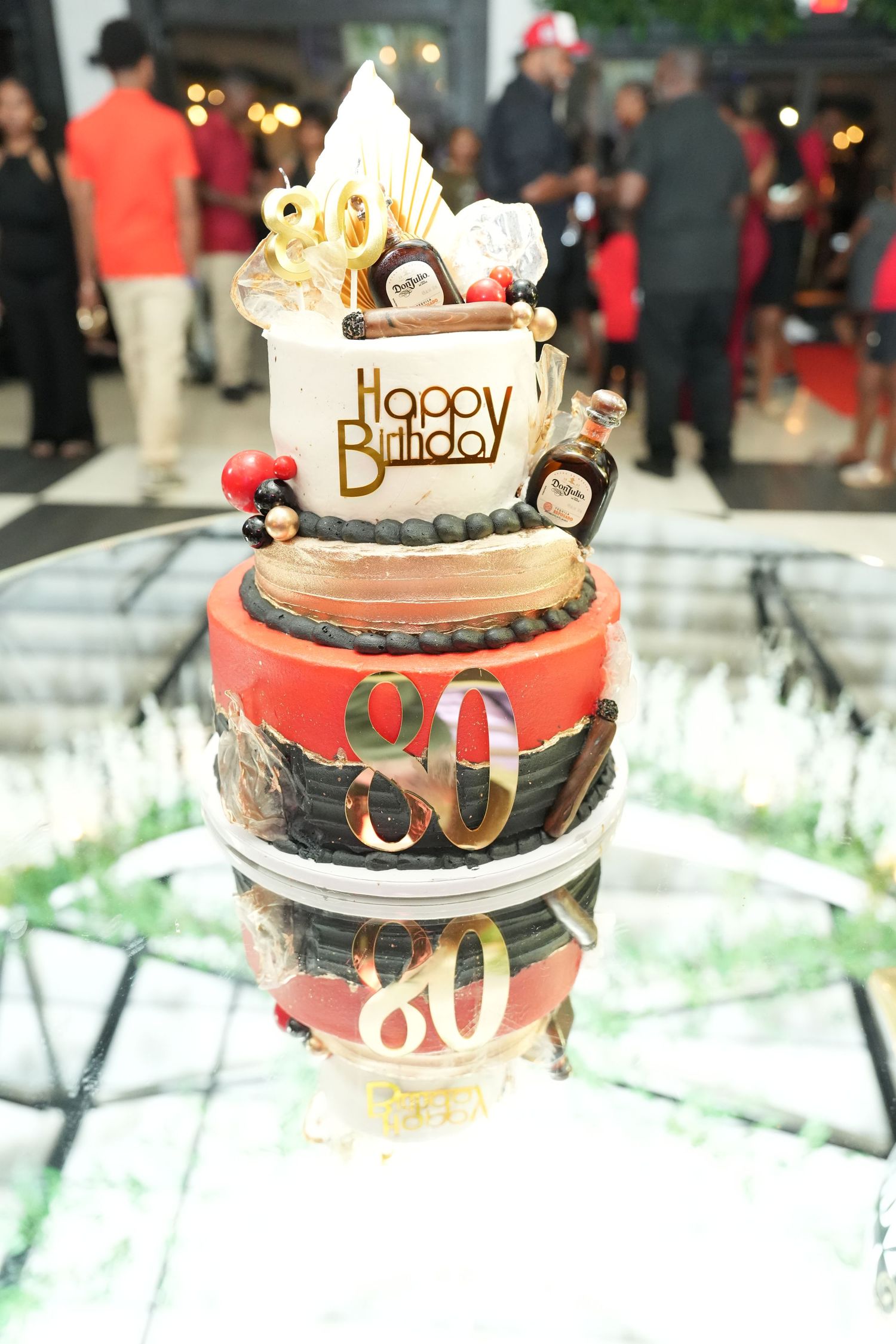 Elegant three-tier 80th birthday cake in red and gold with Happy Birthday topping and decorative liquor bottles.