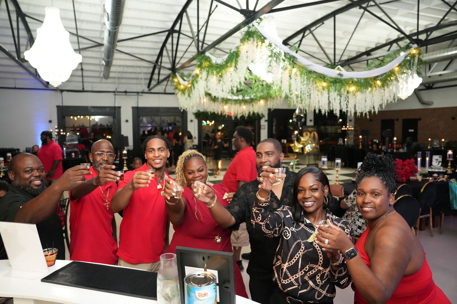 Group of friends in red outfits toasting drinks at a festive indoor party with green floral decorations.