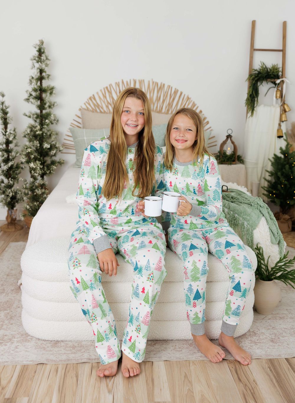 Two girls wearing matching Christmas tree print pajamas sitting on a bed surrounded by holiday decorations.