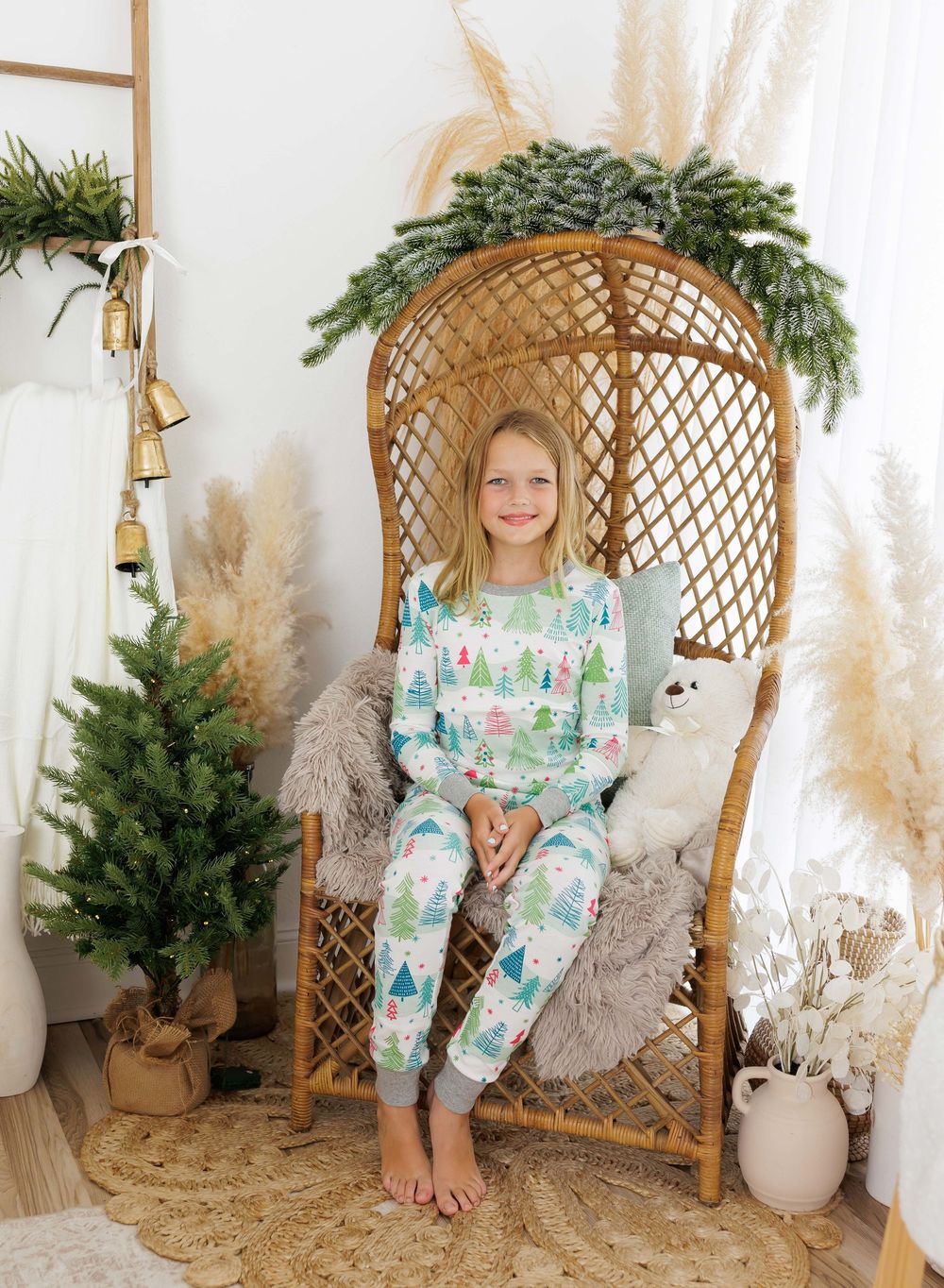 Girl wearing Christmas tree print pajamas sitting in a wicker chair with holiday decorations.