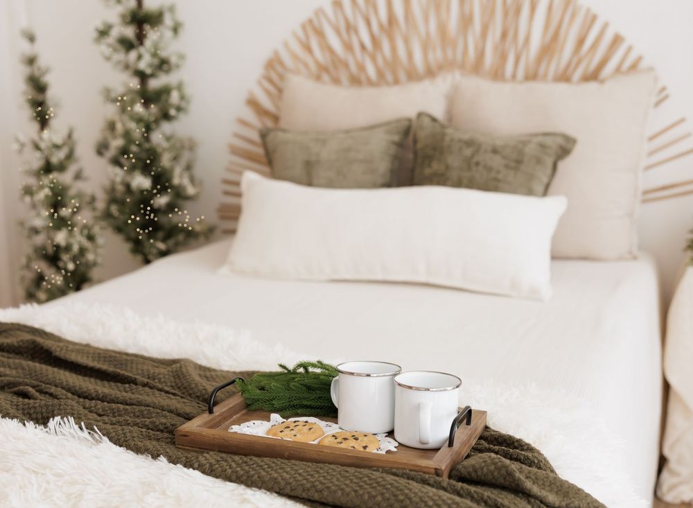 Cozy boho bedroom with white bedding, wooden tray, candles, green throw, and Christmas trees for holiday decor.