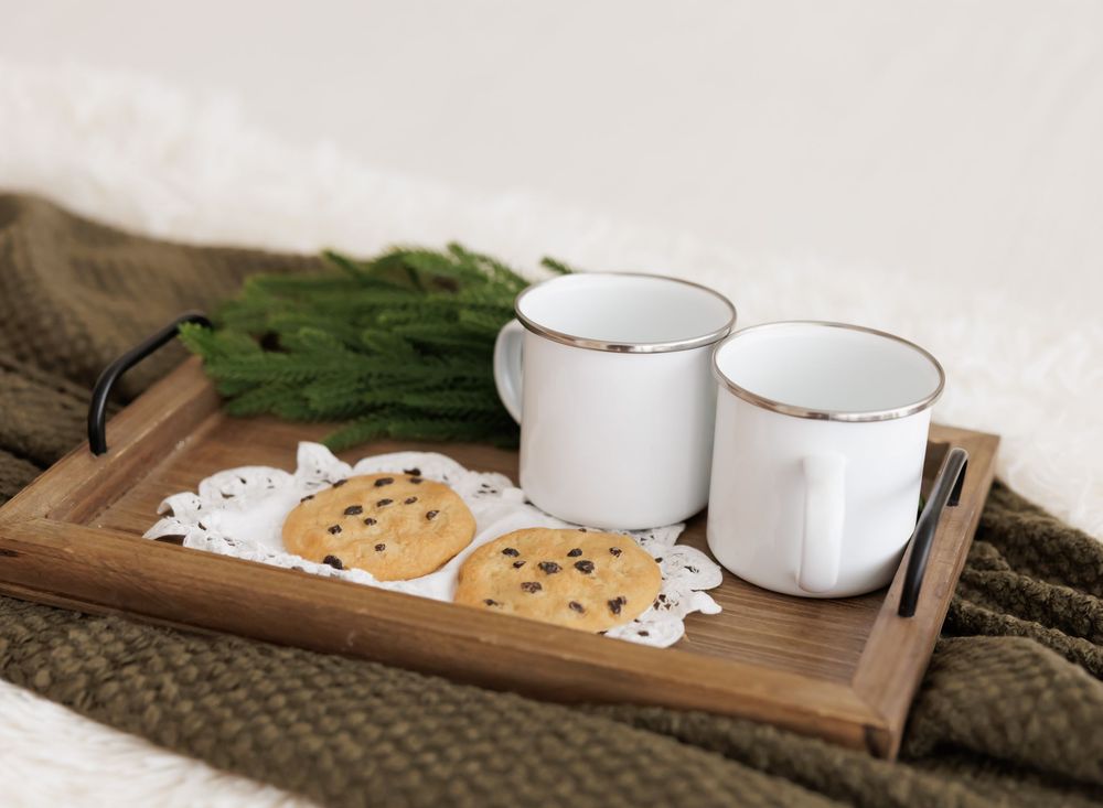 Wooden tray with two white enamel mugs and chocolate chip cookies on doilies, set on a cozy knit blanket.