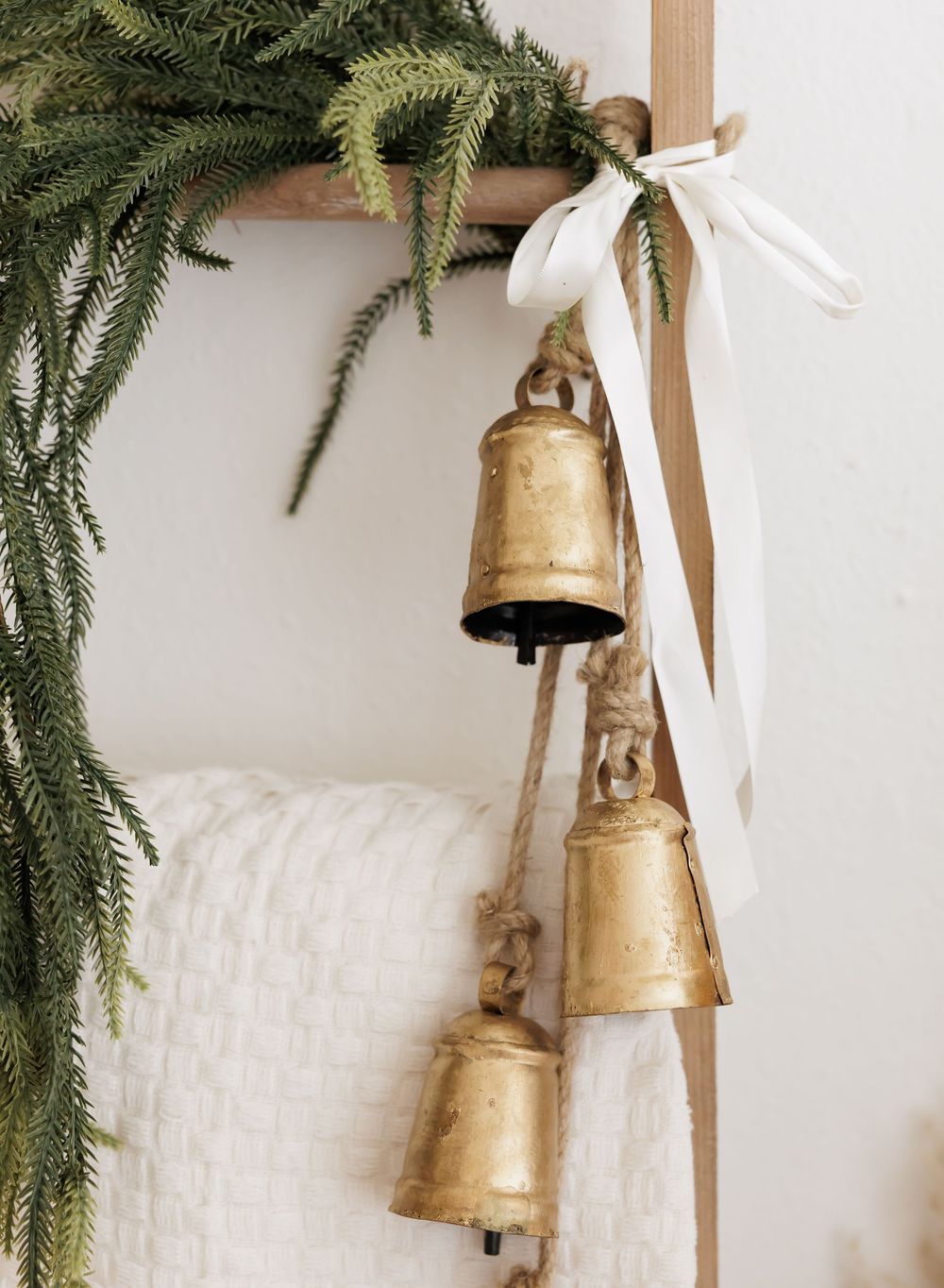 Three rustic gold brass bells hanging from a white ribbon on a wooden ladder with fresh evergreen garland.
