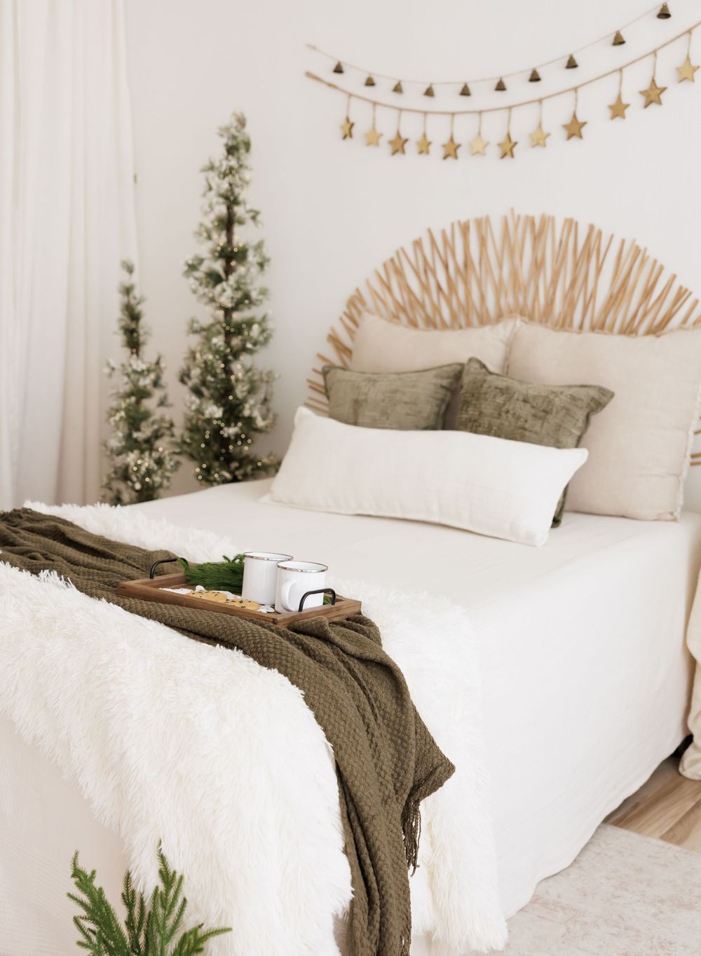 Cozy boho bedroom with white bedding, rattan headboard, green throw, Christmas trees, and gold star garland.