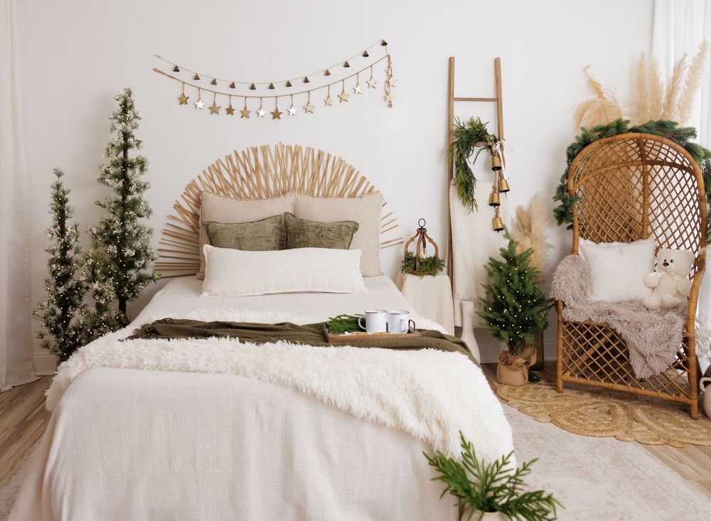 Boho bedroom with white bedding, macrame headboard, Christmas trees, and rattan chair in a cozy holiday setting.