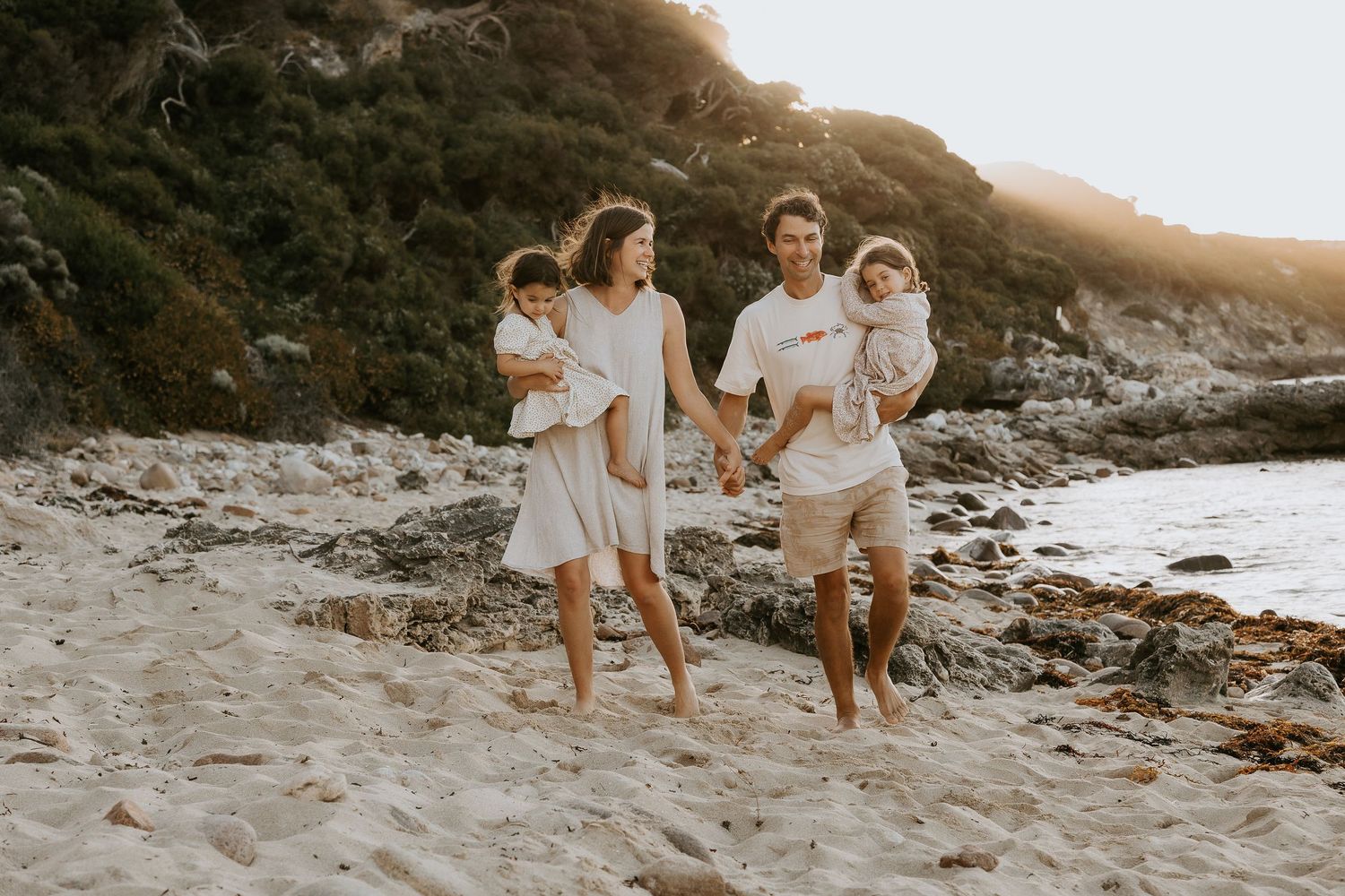 Happy family of four sitting together on a rocky beach shore, smiling warmly during a golden hour photo session.