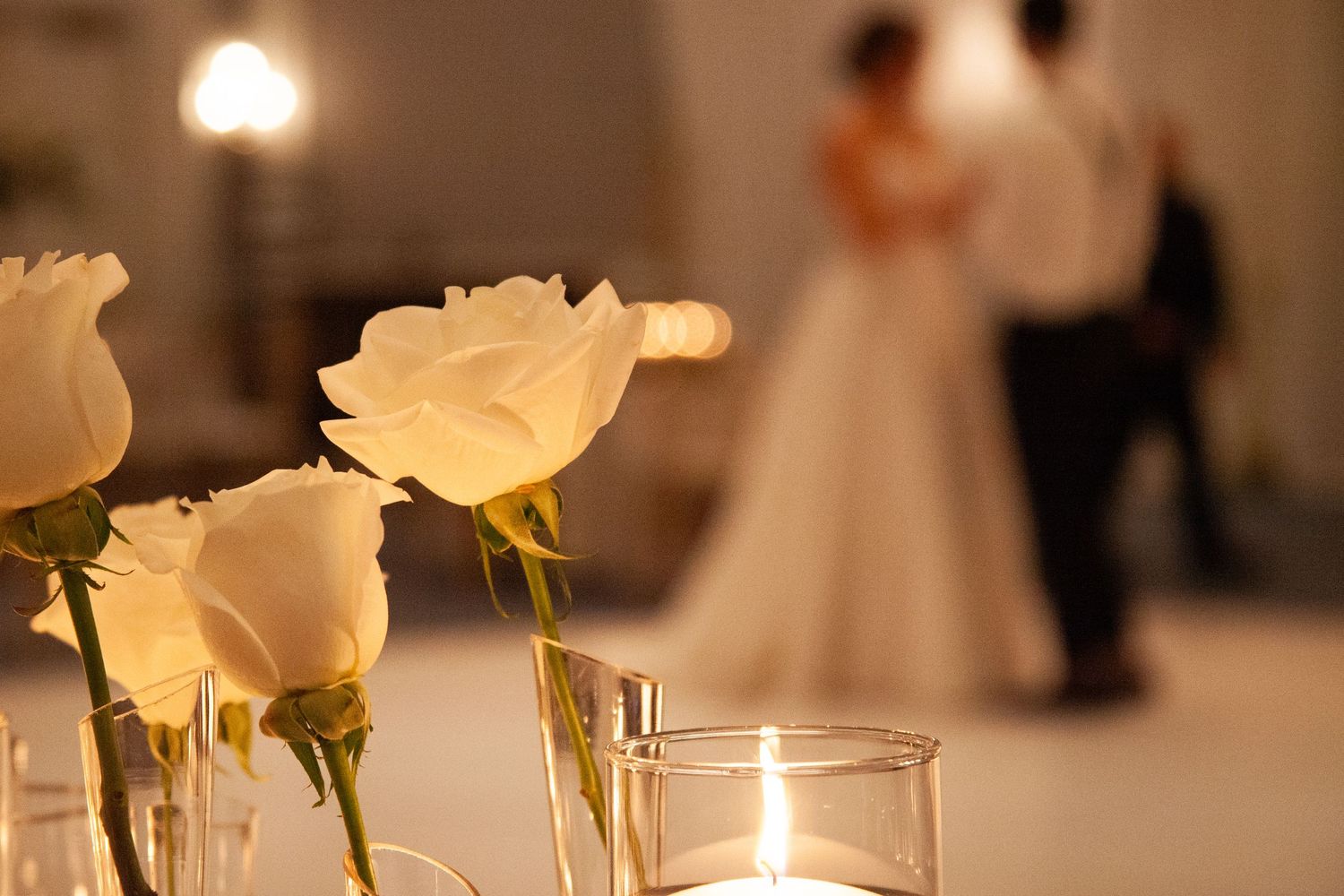 White roses and candles in foreground with couple dancing at romantic wedding reception.