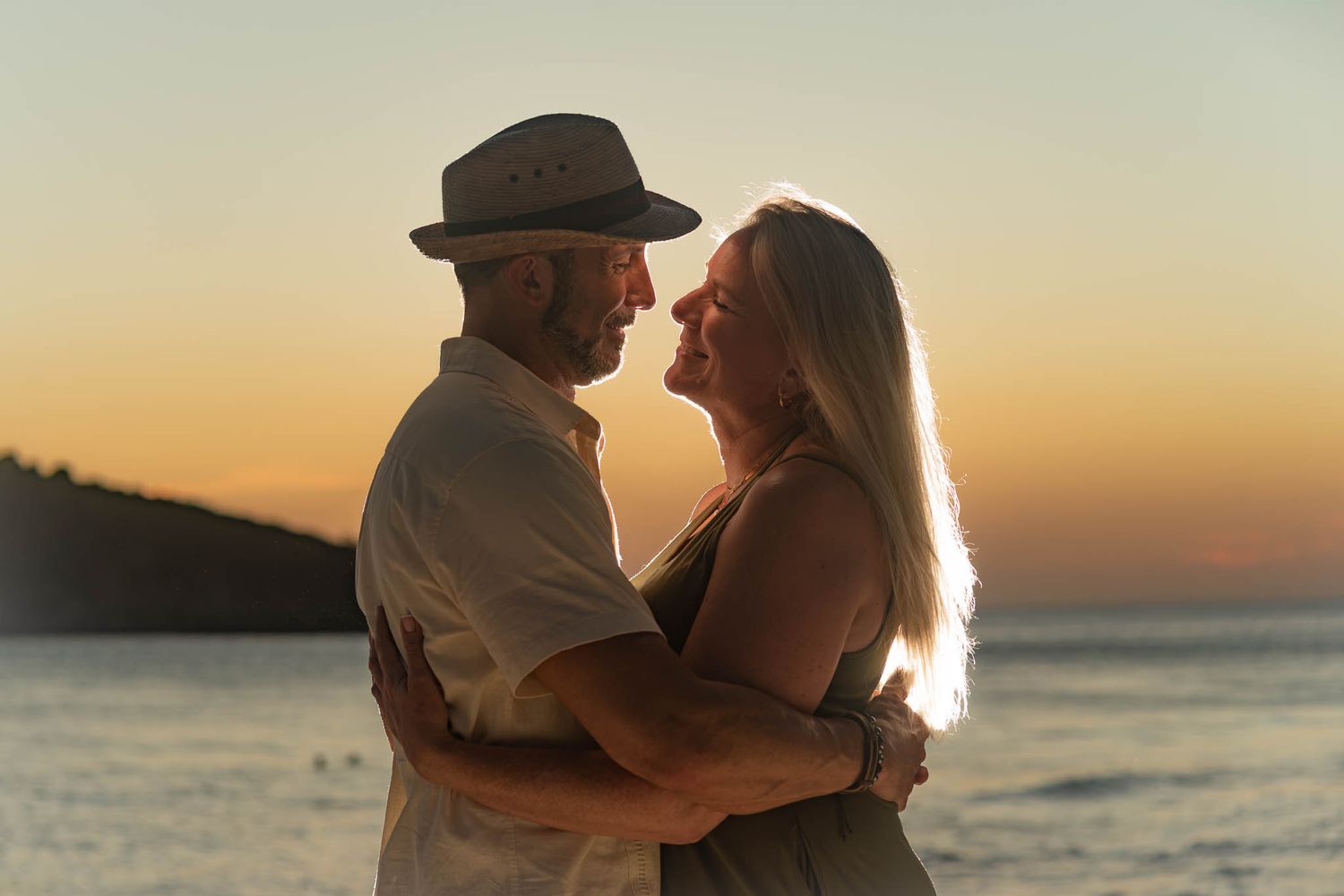 Older couple embracing romantically on a beach at sunset with golden sky and ocean in the background.