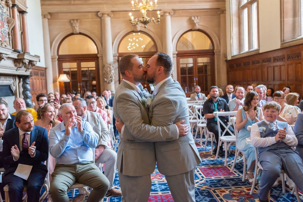 Two grooms share a first kiss at their wedding ceremony in an elegant venue filled with cheering guests.