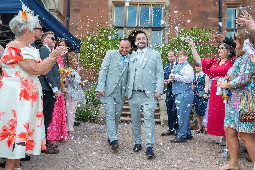 Two grooms in matching grey suits walk through confetti shower surrounded by cheering wedding guests outside a venue.
