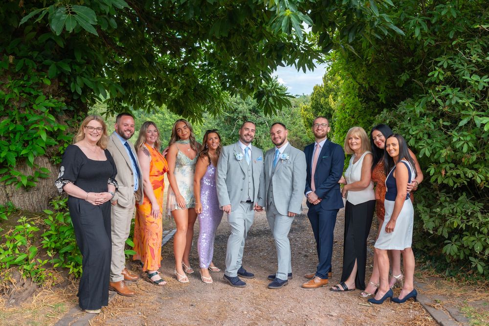 Group of elegantly dressed people posing together on a garden path surrounded by lush green trees.