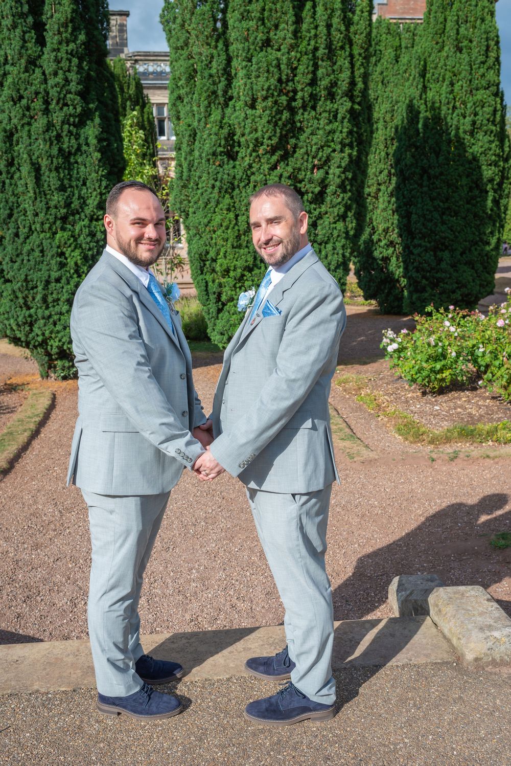 Two grooms in matching grey suits hold hands in a garden setting on their wedding day.