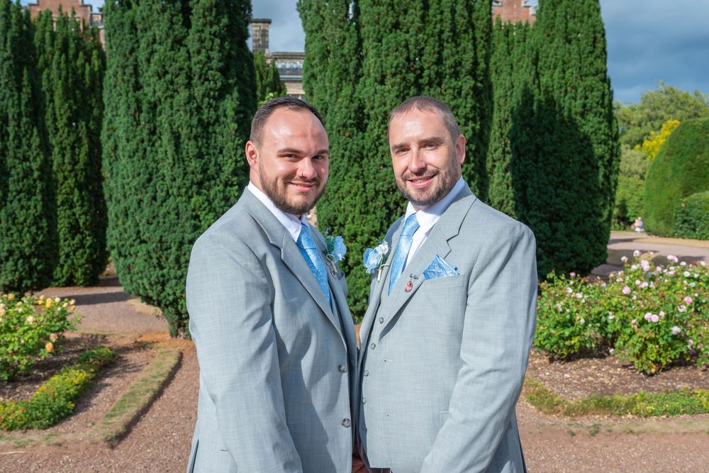 Two men in matching light grey suits with blue boutonnieres pose together in a formal garden with tall cypress trees.